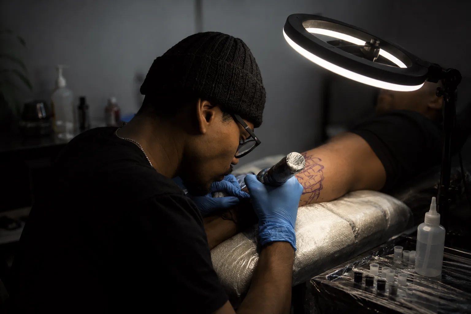 Samuca tatuando um cliente sob a luz do ring light do estúdio — atmosfera reservada, foco no detalhe da arte na pele